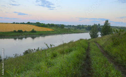 Serene landscape with dirt country road,river and field of ripe wheat.Twilight,early summer morning. 