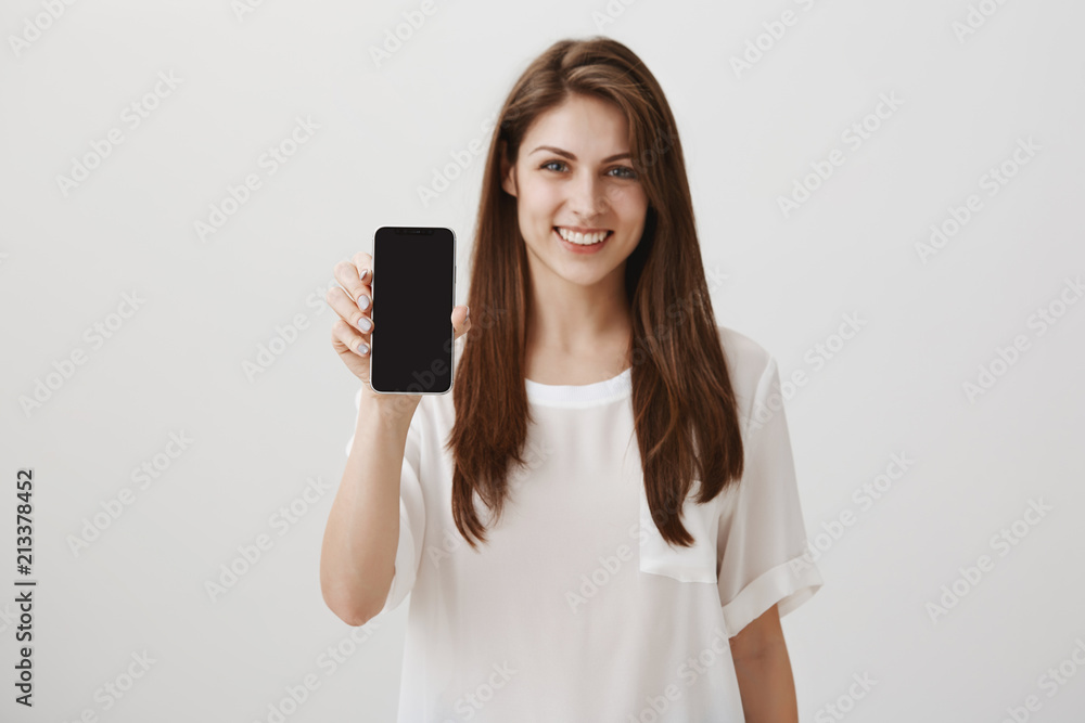 Great device for good price. Portrait of attractive caucasian brunette in white shirt holding smartphone and showing it at camera, smiling broadly, advertising gadget near copy space Great device for good price. Portrait of attractive caucasian brunette in white shirt holding smartphone and showing it at camera, smiling broadly, advertising gadget near copy space