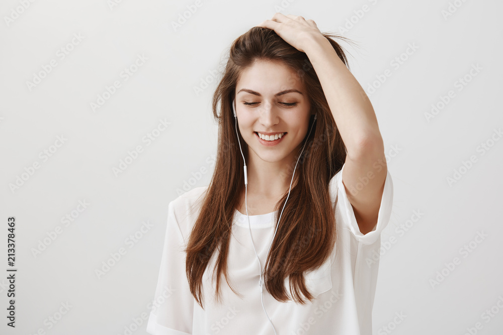 Music that recalls good memories. Portrait of attractive playful woman in white shirt touching hair and looking down with broad smile while listening music in headphones, standing over gray wall Music that recalls good memories. Portrait of attractive playful woman in white shirt touching hair and looking down with broad smile while listening music in headphones, standing over gray wall