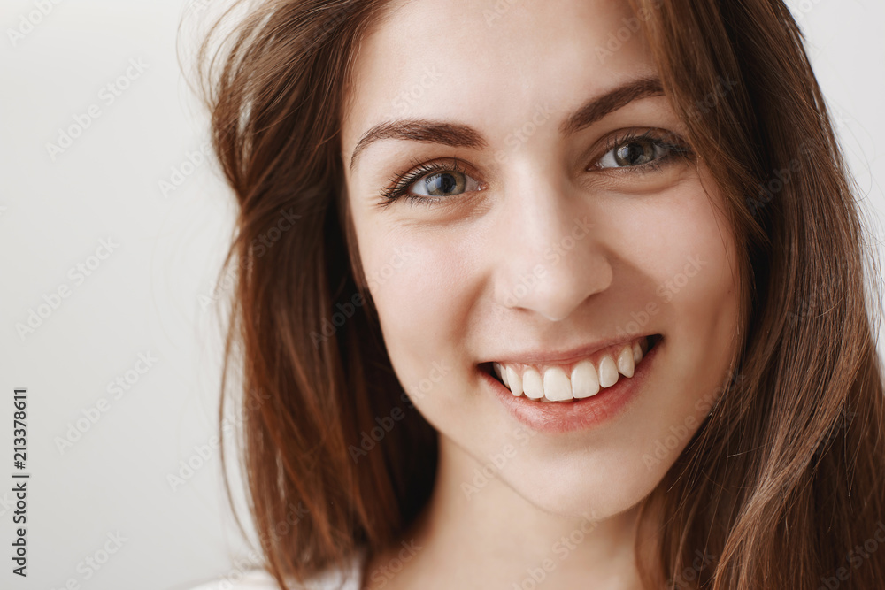 Close-up studio shot of beautiful caucasian brunette smiling broadly and looking with confidence and happiness at camera, standing against gray background, enjoying spending time with colleagues Close-up studio shot of beautiful caucasian brunette smiling broadly and looking with confidence and happiness at camera, standing against gray background, enjoying spending time with colleagues
