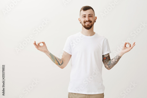 Chrming smile blows mind of every girl. Handsome confident friendly man in white t-shirt, spreading hands and showing zen sign, practicing maditation or yoga, trying something new after busy day photo