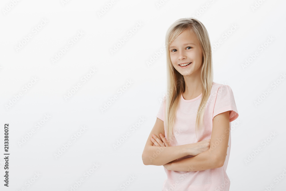 Portrait of smart creative little girl with fair hair, holding hands crossed and smiling broadly, waiting for friend to play on playground together, feeling confident and carefree over gray wall Portrait of smart creative little girl with fair hair, holding hands crossed and smiling broadly, waiting for friend to play on playground together, feeling confident and carefree over gray wall