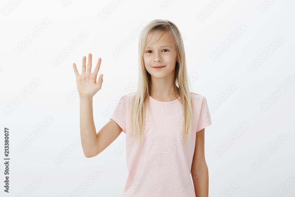 Nice to meet you. Studio shot of friendly smart young caucasian girl with fair hair, raising palm and waving at camera, greeting friend or welcoming classmate, standing over gray wall with cute smile Nice to meet you. Studio shot of friendly smart young caucasian girl with fair hair, raising palm and waving at camera, greeting friend or welcoming classmate, standing over gray wall with cute smile