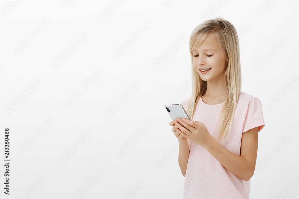 Half-turned profile shot of smart creative female child with blond hair in pink t-shirt, holding smartphone and smiling at screen, playing funny game on device, enjoying spending time over gray wall Half-turned profile shot of smart creative female child with blond hair in pink t-shirt, holding smartphone and smiling at screen, playing funny game on device, enjoying spending time over gray wall