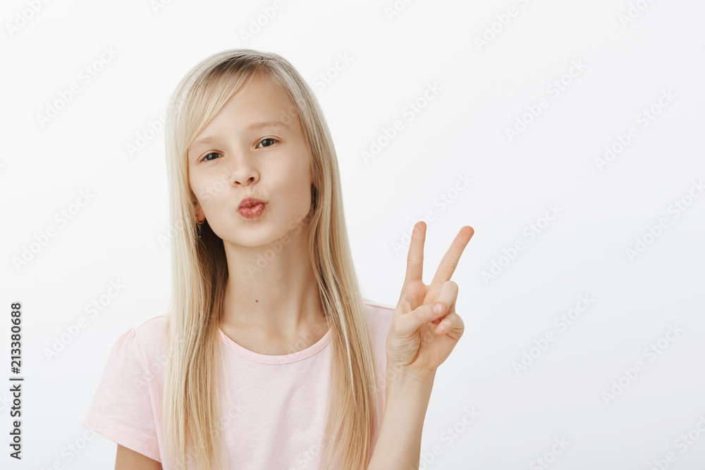 Mwah to my followers, stay updated. Portrait of fashionable confident young girl with natural fair hair, folding lips, blowing kiss, showing peace or victory sign, being in great mood over gray wall Mwah to my followers, stay updated. Portrait of fashionable confident young girl with natural fair hair, folding lips, blowing kiss, showing peace or victory sign, being in great mood over gray wall