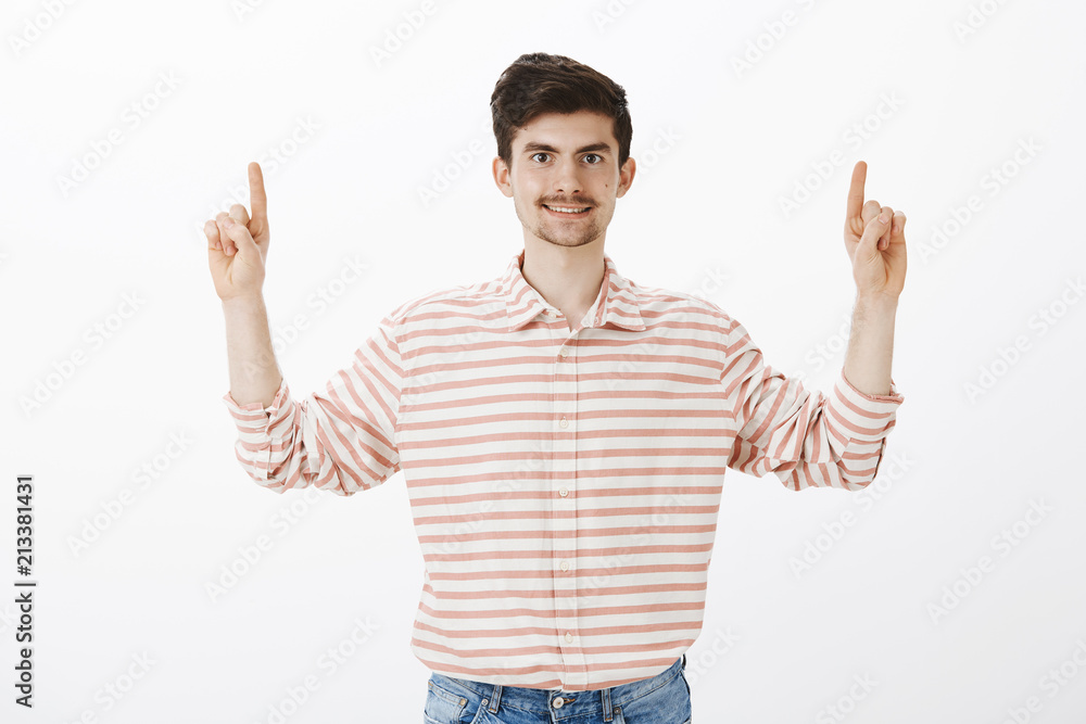 Studio shot of pleased friendly-looking ordinary guy in striped shirt, raising index fingers and pointing up, smiling broadly, suggesting friends to sit in cafe upstairs, standing over gray background Studio shot of pleased friendly-looking ordinary guy in striped shirt, raising index fingers and pointing up, smiling broadly, suggesting friends to sit in cafe upstairs, standing over gray background