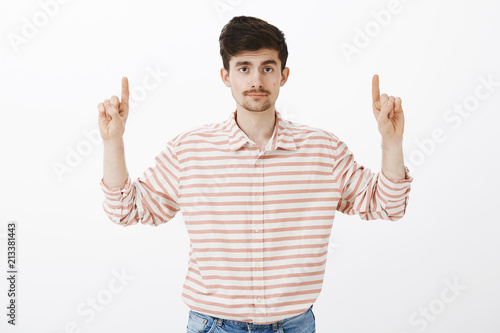 Nothing interesting here. Portrait of indifferent unimpressed handsome guy with beard and moustache, raising hands, pointing up with displeased calm expression, standing against gray background