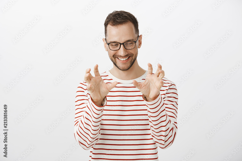 Playful funny european male model in striped pullover and glasses, looking aside while blushing and smiling cheerfully, raising palms and squeezing air as if touching breast, standing over gray wall Playful funny european male model in striped pullover and glasses, looking aside while blushing and smiling cheerfully, raising palms and squeezing air as if touching breast, standing over gray wall