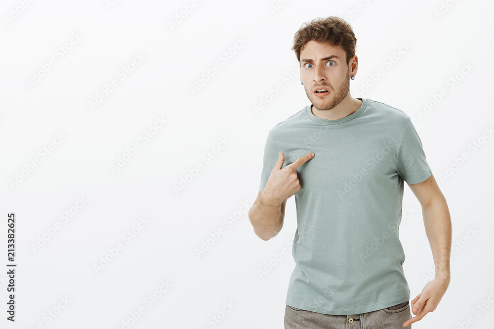 Portrait of shocked frustrated handsome european man in earrings, pointing at himself with index finger and frowning, asking question, being displeased with terrible offencive words over gray wall Portrait of shocked frustrated handsome european man in earrings, pointing at himself with index finger and frowning, asking question, being displeased with terrible offencive words over gray wall