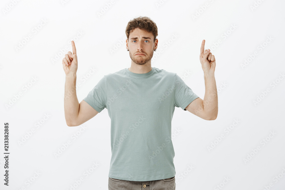 Studio shot of upset miserable handsome man in trendy outfit, raising index fingers and pointing up, being bored or tired after long day working, standing indifferent over gray background Studio shot of upset miserable handsome man in trendy outfit, raising index fingers and pointing up, being bored or tired after long day working, standing indifferent over gray background
