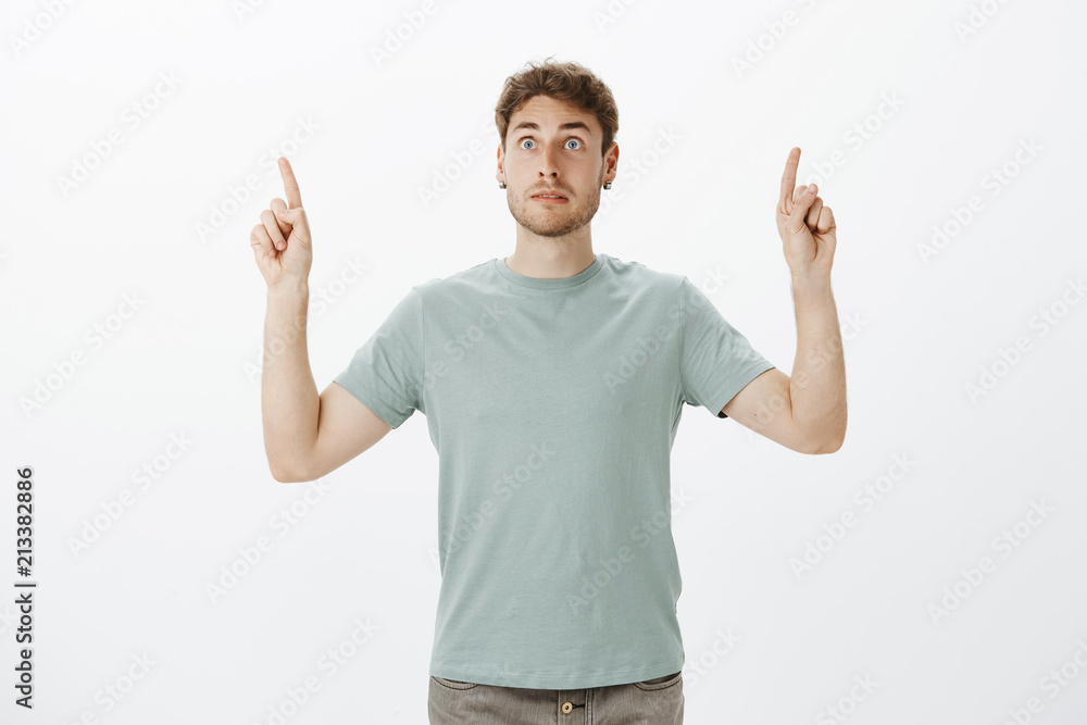 Intense afraid european guy with fair hair, pointing upwards with raised index fingers and staring aside on something scary and shocking, being stunned and speechless over gray background Intense afraid european guy with fair hair, pointing upwards with raised index fingers and staring aside on something scary and shocking, being stunned and speechless over gray background