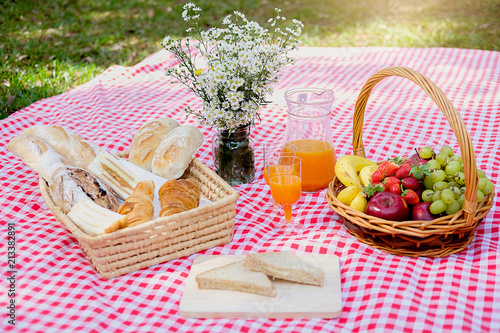 Picnic wicker basket with food, bread, fruit and orange juice on a red and white checked cloth in the field with green nature background. Picnic concept.