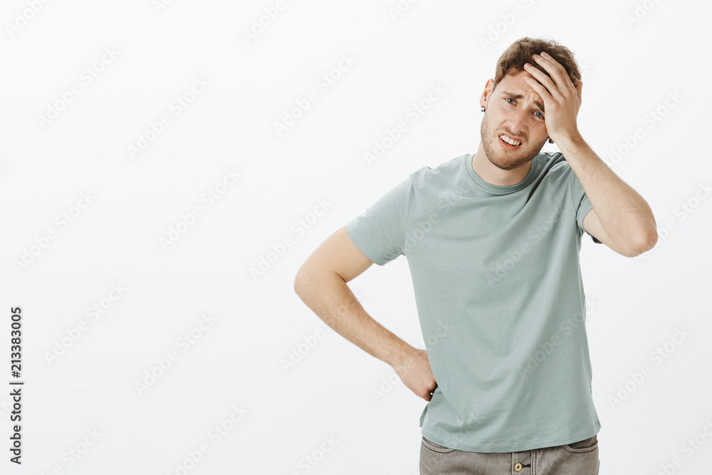 Portrait of troubled gloomy european male model in casual outfit, holding palm on forehead and frowning, touching hip with arm, feeling headache from overworking, facing problems in life Portrait of troubled gloomy european male model in casual outfit, holding palm on forehead and frowning, touching hip with arm, feeling headache from overworking, facing problems in life