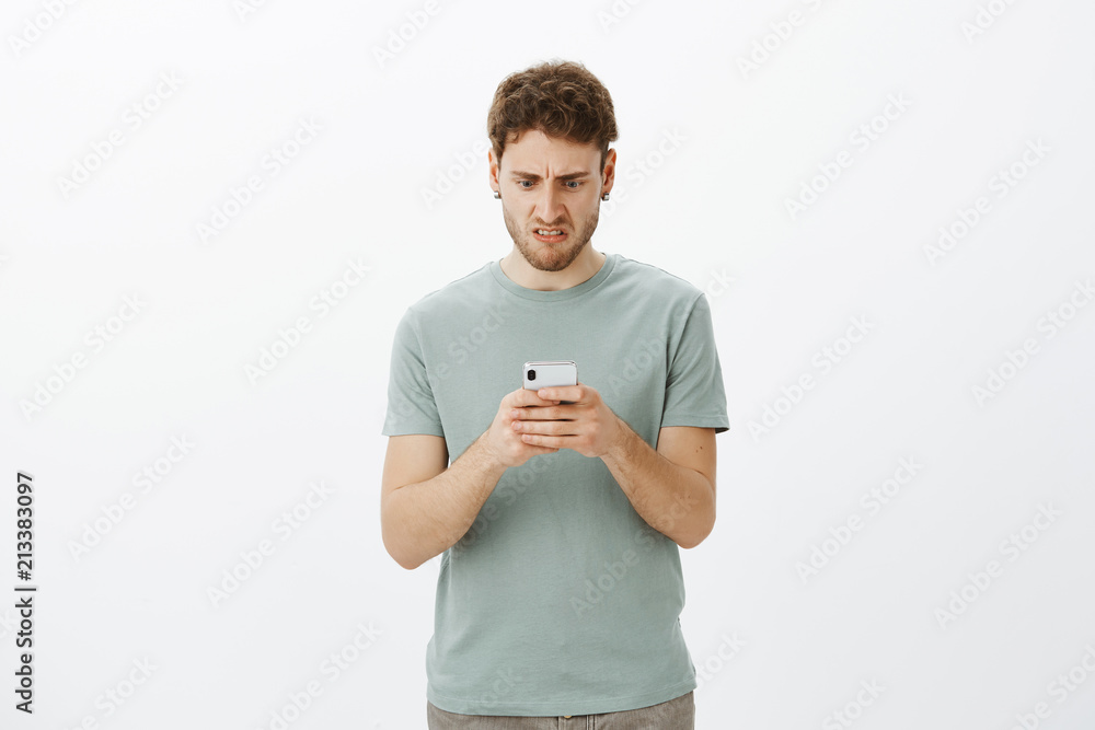 Frustrated disgusted Caucasian man, holding smartphone and staring displeased at screen, grimacing from negative emotions, disliking new article he read in device, standing against gray wall Frustrated disgusted Caucasian man, holding smartphone and staring displeased at screen, grimacing from negative emotions, disliking new article he read in device, standing against gray wall