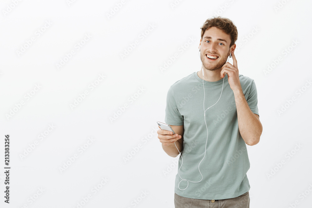 Studio shot of satisfied pleased caucasian male model with bristle, holding smartphone and touching earphone while listening music and enjoying nice sound of earbuds, standing happy over grey wall Studio shot of satisfied pleased caucasian male model with bristle, holding smartphone and touching earphone while listening music and enjoying nice sound of earbuds, standing happy over grey wall