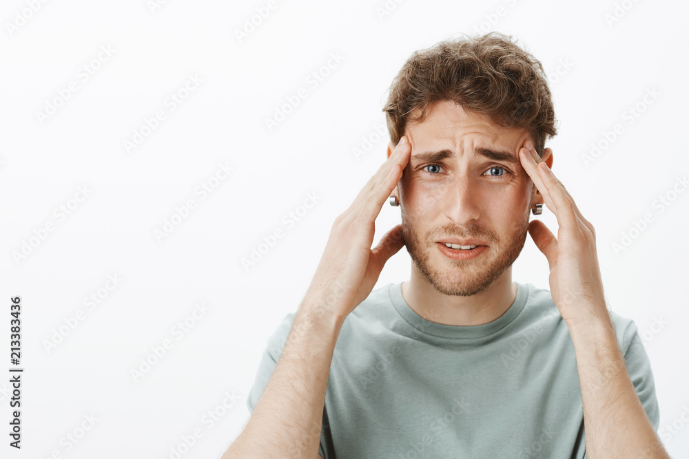 Close-up shot of troubled displeased unhappy male model with fair hair, holding hands on temples and frowning, feeling pain or migraine, suffering from headache over gray background Close-up shot of troubled displeased unhappy male model with fair hair, holding hands on temples and frowning, feeling pain or migraine, suffering from headache over gray background