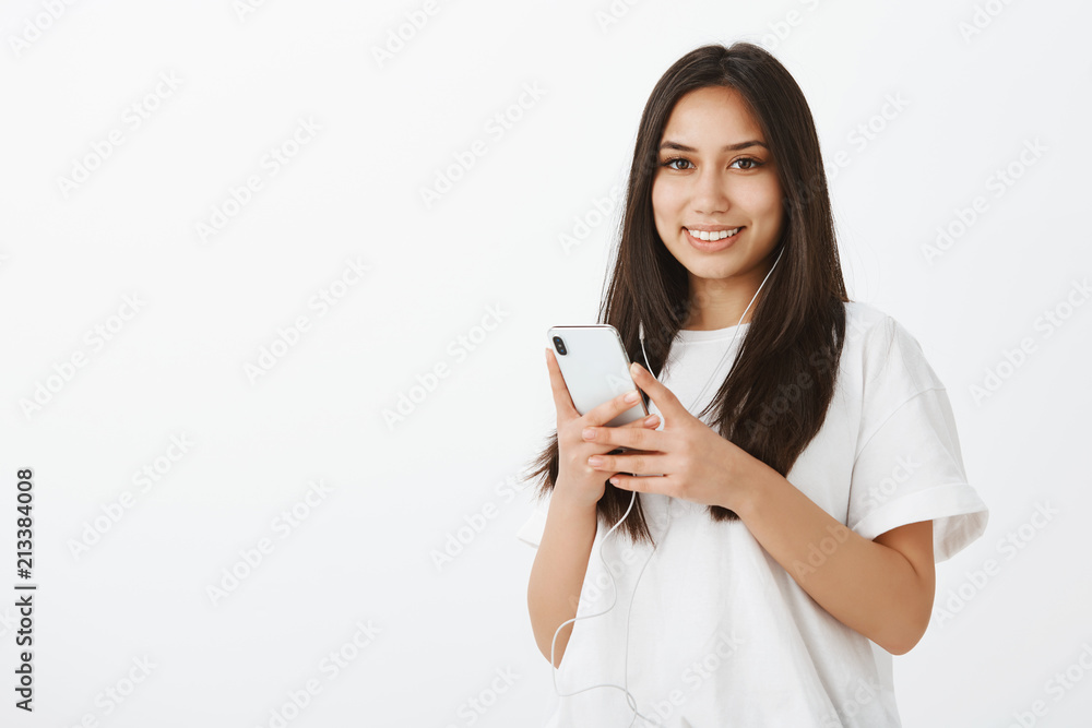 Happy beautiful Caucasian girl in white t-shirt, holding smartphone and listening music in earphones, gazing with cheerful smile at camera, picking track to go for walk, standing over gray wall Happy beautiful Caucasian girl in white t-shirt, holding smartphone and listening music in earphones, gazing with cheerful smile at camera, picking track to go for walk, standing over gray wall