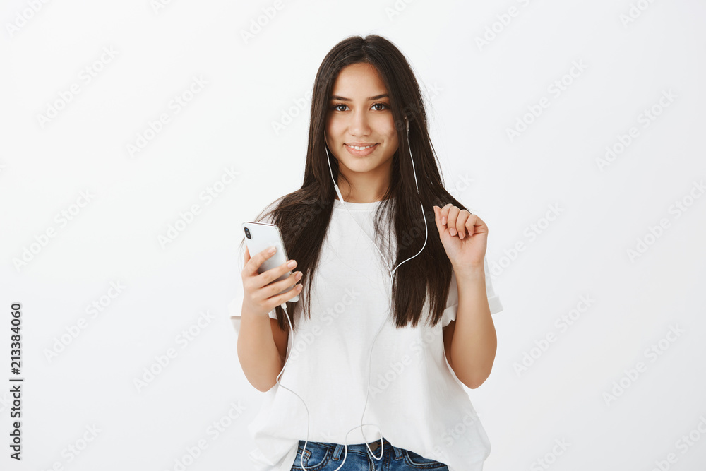 Portrait of flirty attractive feminine woman in white t-shirt, raising hand and holding smartphone while listening music in earphones and gazing at camera, feeling joy and relaxation over gray wall Portrait of flirty attractive feminine woman in white t-shirt, raising hand and holding smartphone while listening music in earphones and gazing at camera, feeling joy and relaxation over gray wall