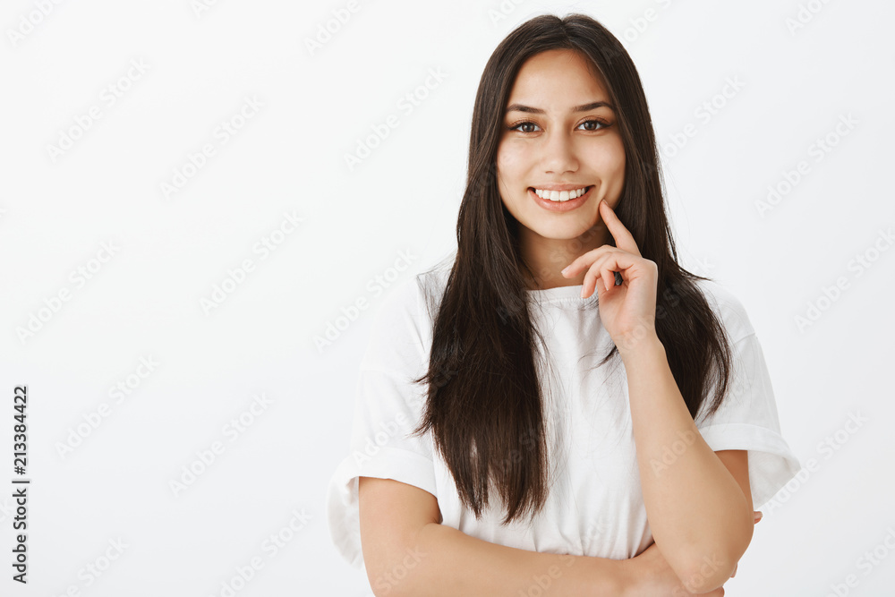 Waist-up shot of confident feminine european woman with tanned skin and dark hair, holding finger on chin and smiling joyfully while gazing at camera, standing against gray background Waist-up shot of confident feminine european woman with tanned skin and dark hair, holding finger on chin and smiling joyfully while gazing at camera, standing against gray background