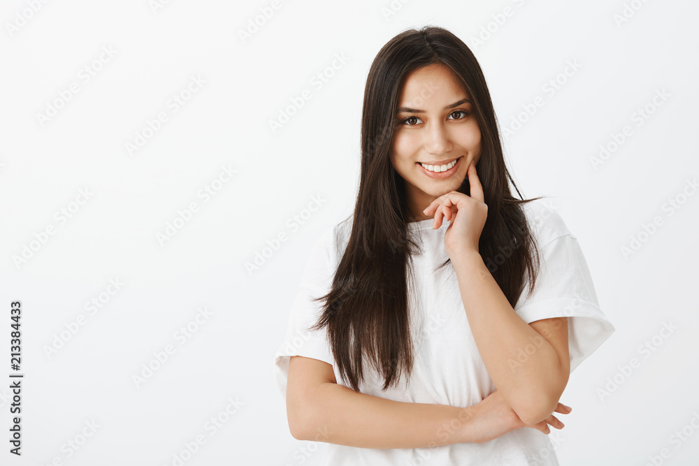 Waist-up portrait of positive friendly-looking girlfriend with dark hair, touching chin with fingers and smiling joyfully, having creative idea or thought, planning perfect date over gray background Waist-up portrait of positive friendly-looking girlfriend with dark hair, touching chin with fingers and smiling joyfully, having creative idea or thought, planning perfect date over gray background