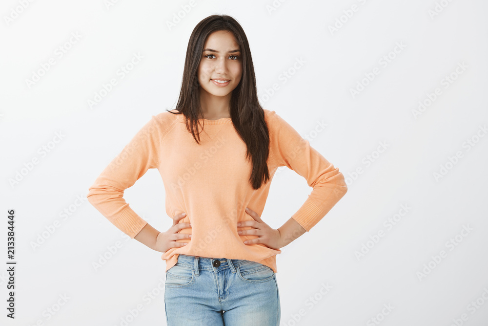 Studio shot of self-assured attractive european girl with dark hair, holding hands on hips and gazing cheerfully at camera with joyful smile, posing for advertisement or during beauty contest Studio shot of self-assured attractive european girl with dark hair, holding hands on hips and gazing cheerfully at camera with joyful smile, posing for advertisement or during beauty contest