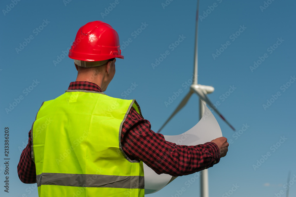 Rear view of unknown male engineer in a green waistcoat with red helmet with engineering plans against the background of windmills and the blue sky Rear view of unknown male engineer in a green waistcoat with red helmet with engineering plans against the background of windmills and the blue sky