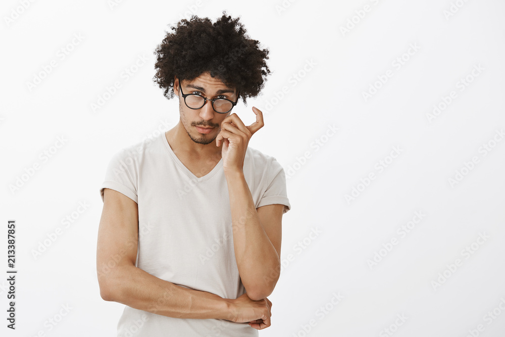 Indoor shot of charming smart hispanic male model with curly hair and moustache in glasses, frowning and gazing with interest from under forehead, scratching cheek, thinking over grey wall Indoor shot of charming smart hispanic male model with curly hair and moustache in glasses, frowning and gazing with interest from under forehead, scratching cheek, thinking over grey wall