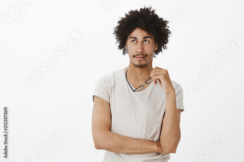 Indoor shot of attractive smart and relaxed young male with afro haircut and moustache, looking up and biting rim of glasses while dreaming or thinking, making plan in mind over gray background