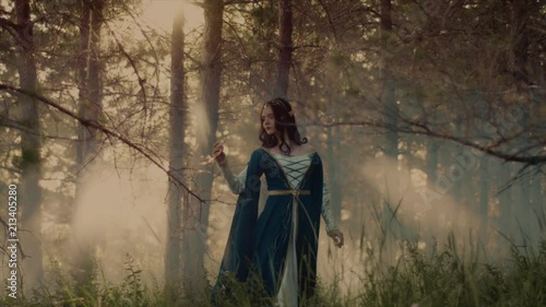 Pretty unusual woman looking like a medieval elf princess is strolling on sunny morning in woodland. She is looking around, enjoying nature and sunrise