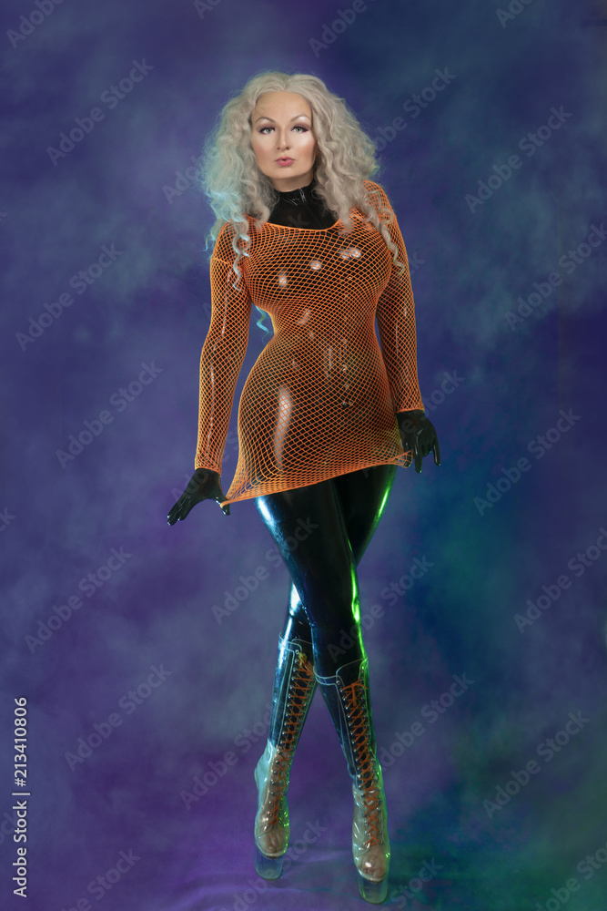 Cute Chubby Adult Female In Black Latex Rubber Catsuit And Orange Mesh Dress Is Standing In
