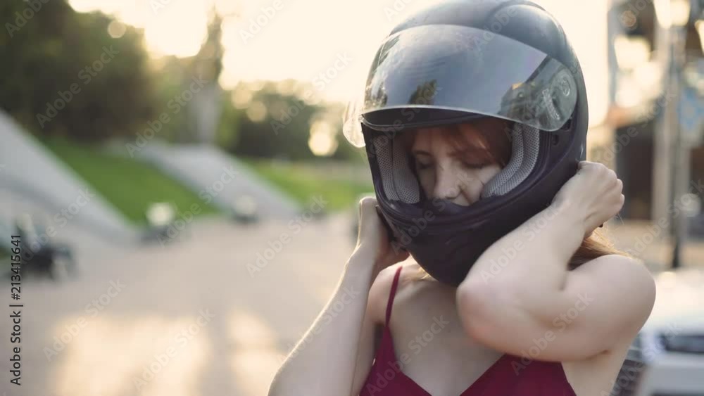Beautiful young red-haired woman motorcyclist dresses her black helmet. On street at sunset or sunrise Beautiful young red-haired woman motorcyclist dresses her black helmet. On street at sunset or sunrise