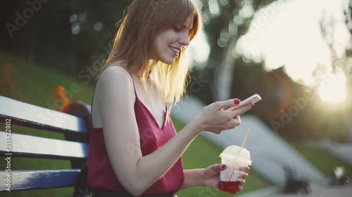 Beautiful young red-haired  woman sitting on a bench in the park drinking juice and using a smartphone on sunset  photo