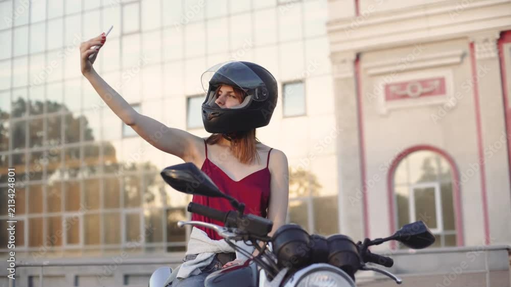 Beautiful young red-haired woman motorcyclist with her motorcycle and helmet. On street at sunset or sunrise. Woman biker Beautiful young red-haired woman motorcyclist with her motorcycle and helmet. On street at sunset or sunrise. Woman biker