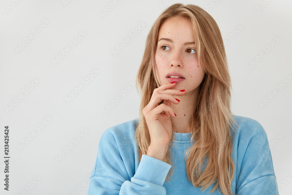 Pensive European female holds hand on chin, looks aside with dreamy expression, contemplates about someting, dressed casually, isolated over white blank wall with copy space aside for promotion Pensive European female holds hand on chin, looks aside with dreamy expression, contemplates about someting, dressed casually, isolated over white blank wall with copy space aside for promotion