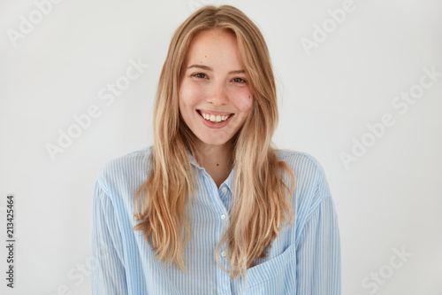 Cheerful blonde young female with attractive look, broad shining smile, dressed in fashionable shirt, stands against white background, being in high spirit after stroll during sunny spring day © wayhome.studio 