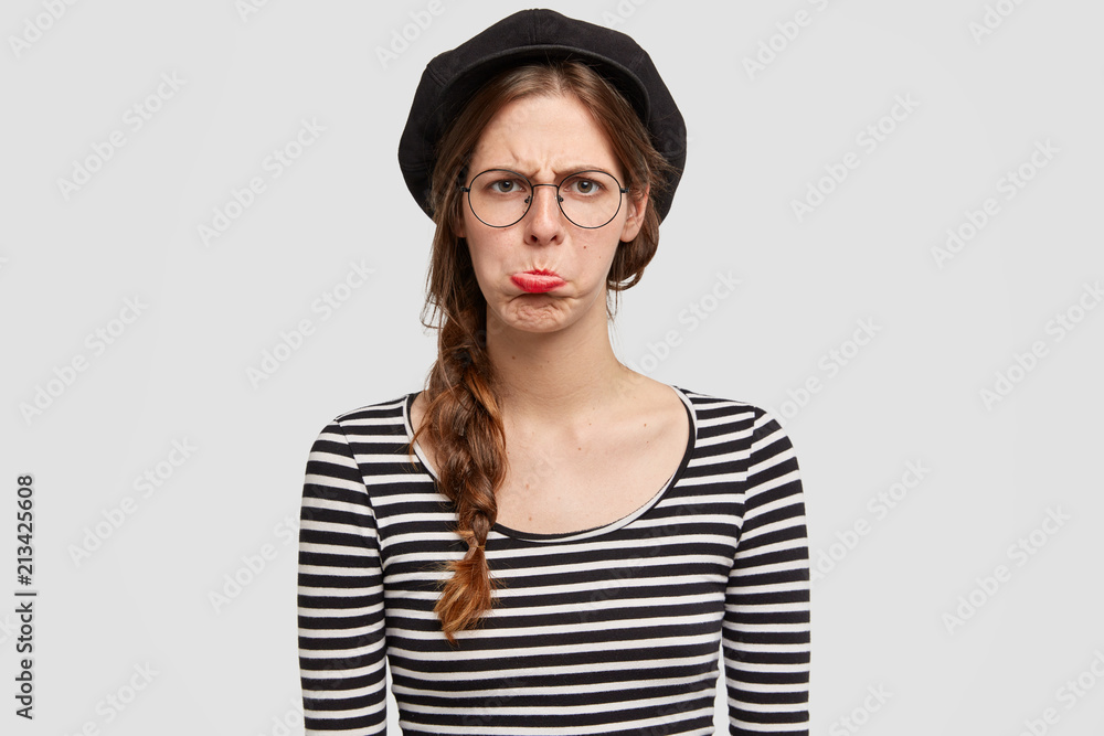 People and insult concept. Beautiful young Parisian female being abused by negative news, purses lower lip, wears casual clothes in French style, isolated over white background, has problems in life People and insult concept. Beautiful young Parisian female being abused by negative news, purses lower lip, wears casual clothes in French style, isolated over white background, has problems in life