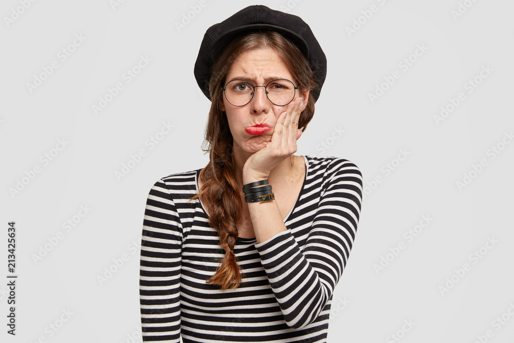 Discontent Parisian female purses lower lip and touches cheek, has unhappy sorrowful expression, displeased with relationship, has truly French style in appearance and clothing, feels insult Discontent Parisian female purses lower lip and touches cheek, has unhappy sorrowful expression, displeased with relationship, has truly French style in appearance and clothing, feels insult