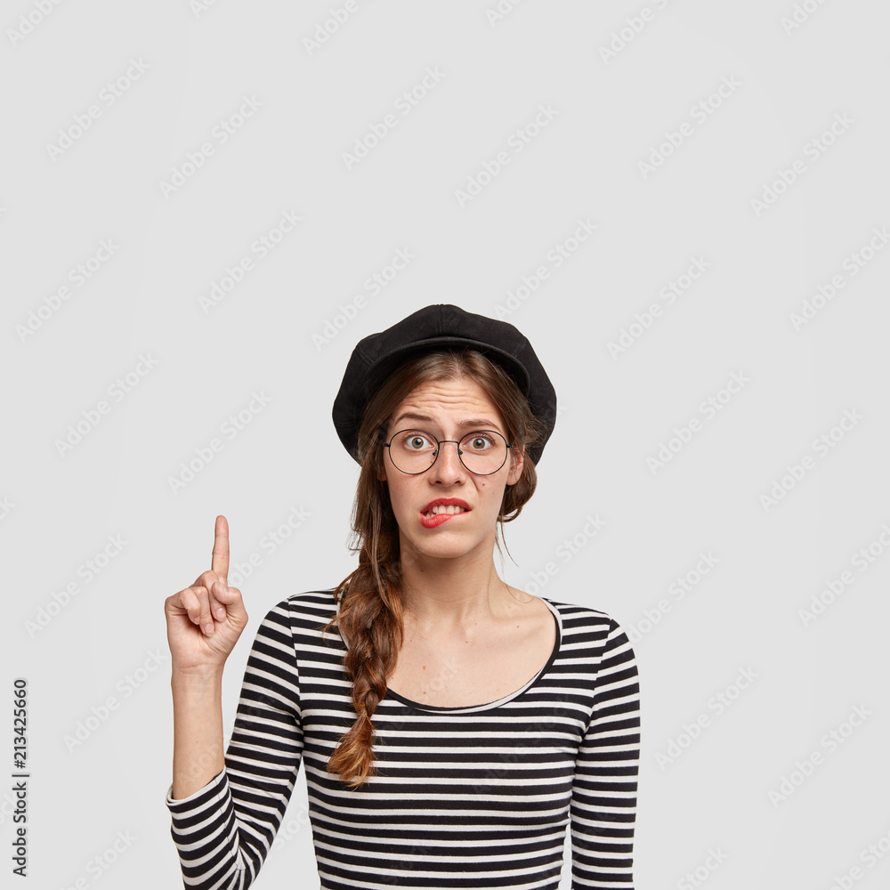 Hesitant Parisian female bites lower lip in puzzlement, points upwards with index finger, feels clueless whether to buy something, dressed in striped sweater, isolated over white background. Hesitant Parisian female bites lower lip in puzzlement, points upwards with index finger, feels clueless whether to buy something, dressed in striped sweater, isolated over white background.