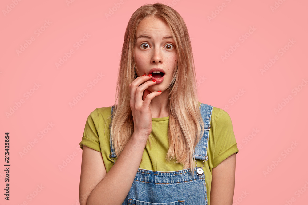 Indoor shot of beautiful young female with shocked expression, opens eyes and mouth widely, hears surprising news, wears denin overalls, stands against pink background. People, emotions concept Indoor shot of beautiful young female with shocked expression, opens eyes and mouth widely, hears surprising news, wears denin overalls, stands against pink background. People, emotions concept