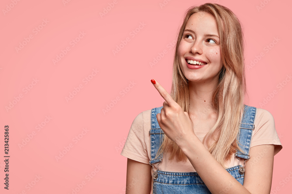 Adorable cheerful young Caucasian female with positive smile, points aside, shows blank space, being in high spirit, dressed in casual clothes, isolated over pink background. Look aside, please! Adorable cheerful young Caucasian female with positive smile, points aside, shows blank space, being in high spirit, dressed in casual clothes, isolated over pink background. Look aside, please!