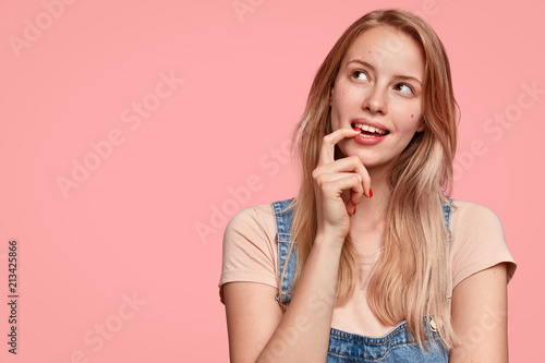 Dreamy Caucasian female with thoughtful expression, looks away, keeps hand near face, thinks about something pleasant, poses against pink background with blank copy space for your promotion.
