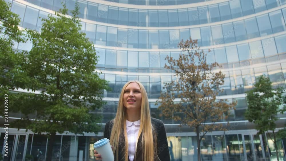 Stylish business woman with coffee on street. From below of serious blond executive woman in suit having coffee in paper cup looking away on street. Stylish business woman with coffee on street. From below of serious blond executive woman in suit having coffee in paper cup looking away on street.