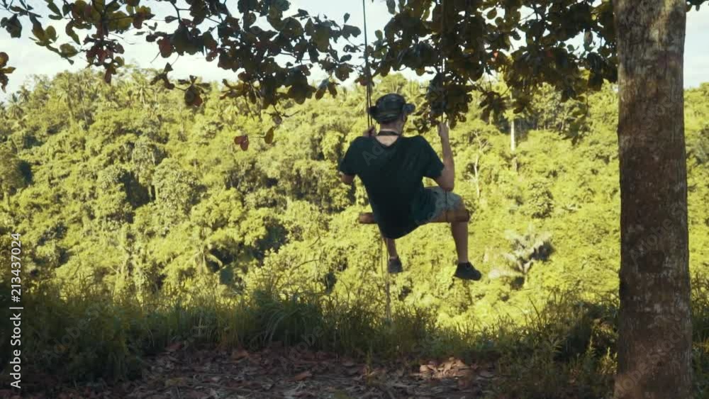 Young male tourist swings on a rope swing hanging from a tree over looking a valley Young male tourist swings on a rope swing hanging from a tree over looking a valley