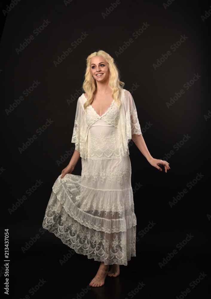 full length portrait of blonde girl wearing white lace dress. standing pose. black studio background.