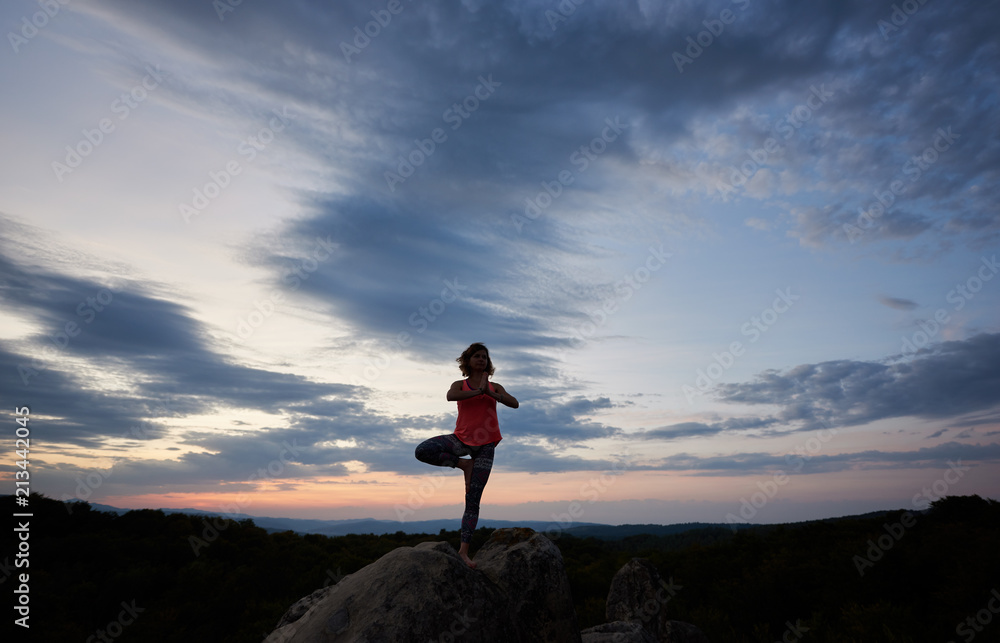 Obraz premium Full-length picture of young slim fit woman doing yoga exercises standing barefooted on one leg on big mountain rock against beautiful evening sky. Fitness and active lifestyle concept. Vrikshasana