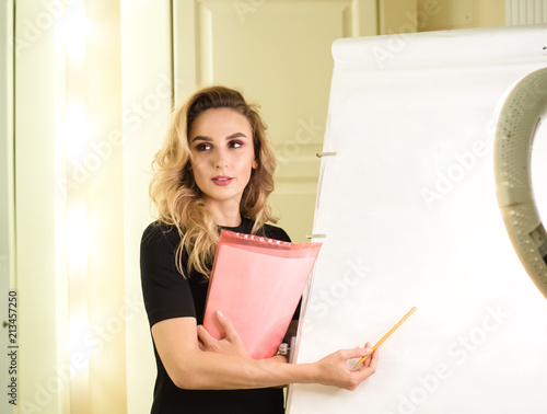 Pretty girl with curly hair standing with folders. Reporter with files next to white board. Reporter, lecture, course, classes teacher. Presentation, studying, report, business concept