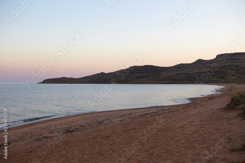 Deserted sandy beach. Peaceful bay with rocky coastline at sunset. Sun setting behind hills at seaside. Tranquil sea with blue and pink morning sky. Summertime, seascape, travel and tourism