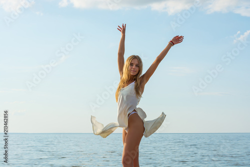 Beautiful happy young woman in a white summer satin dress stands at sea background