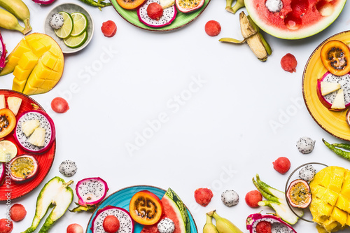 Summer various colorful sliced tropical fruits and berries in plates and bowls on white background, top view, frame. Clean and healthy lifestyle background