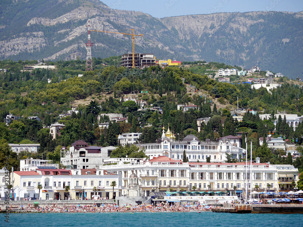The sea embankment with a large number of tourists. With the town houses behind them and the beautiful high mountains.